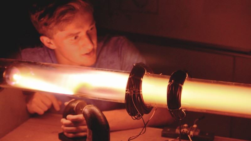 A man kneeling behind a glowing tube holds a magnet to bend the red-orange plasma up in the left of the frame. The tube has two wound electromagnets around it, currently turned off and exerting no effect on the plasma.